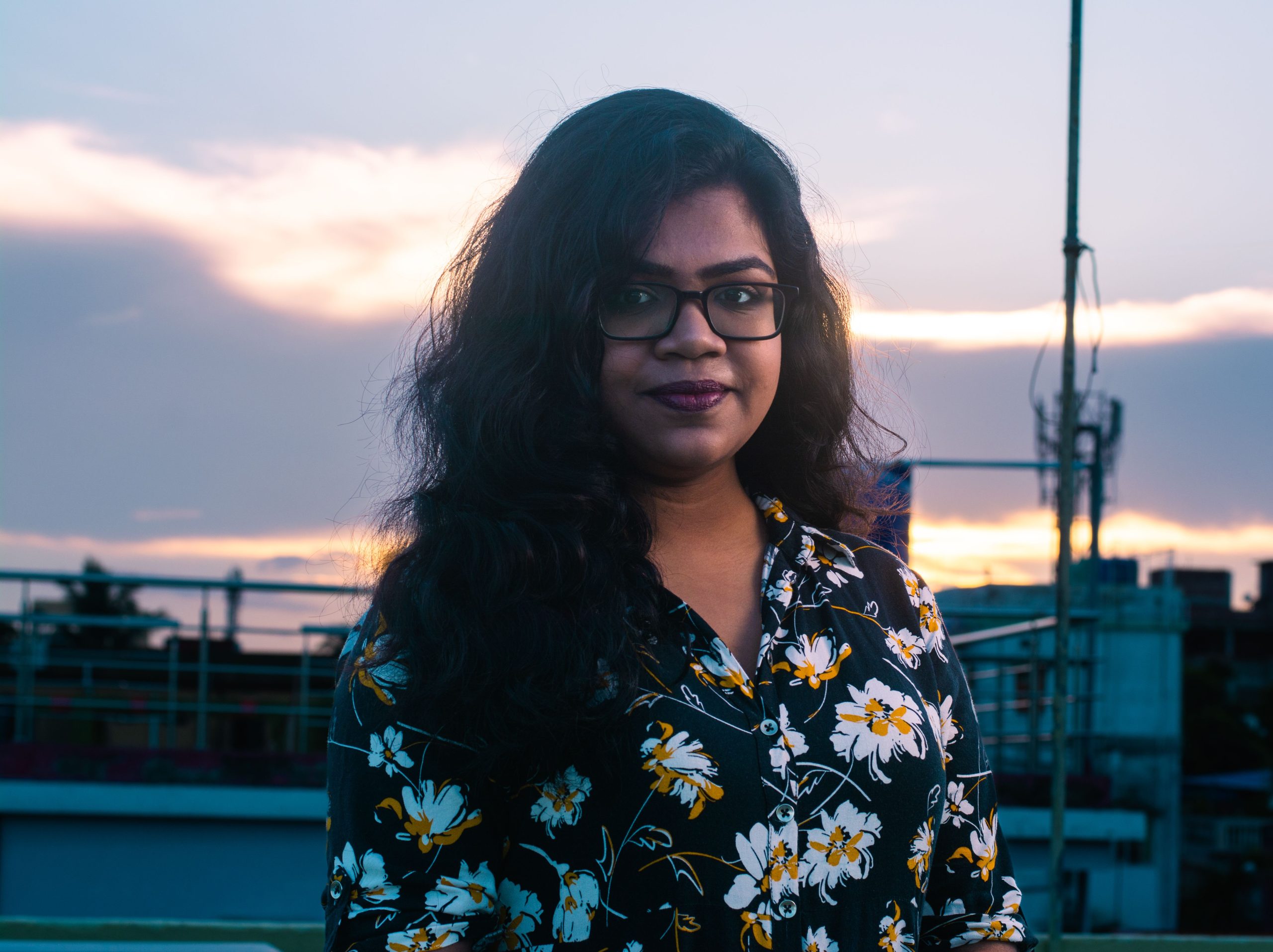 A woman in glasses and floral shirt poses on a rooftop at dusk, with an urban skyline behind her.